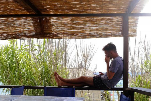 Jovem sentado no gazebo enquanto olha para seu smartphone. No fundo as palhinhas criam uma cena tropical
. - Foto, Imagem