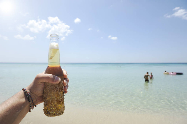 Mannelijke hand houden een fles bier tegen een zonnige hemel en kristalheldere zee. Op de achtergrond hebben sommige mensen plezier in het water. Vakantie concept. - Foto, afbeelding