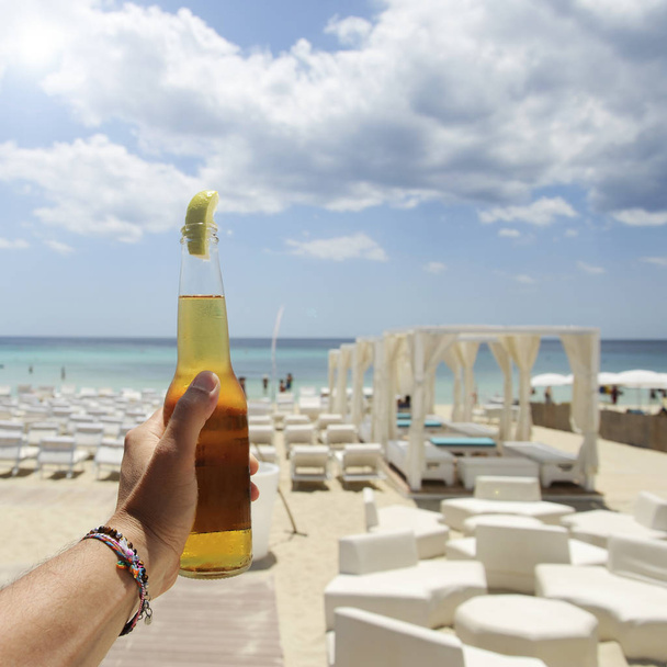Mano masculina sosteniendo una botella de cerveza contra un cielo soleado y un mar cristalino. En el fondo un hermoso complejo de playa. Concepto de vacaciones
. - Foto, imagen