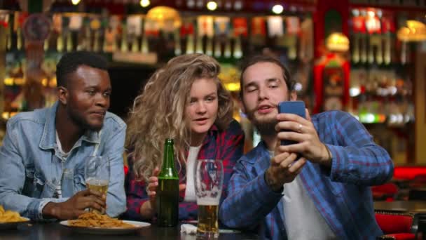 Multi-ethnic group of friends take a selfie in a bar and laugh with a beer while looking at photos on a smartphone screen. - Footage, Video