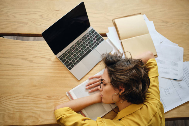 若い疲れた女性のトップビューは、職場で頭の下にノートパソコンやドキュメントで机の上に眠りに落ちる - 写真・画像