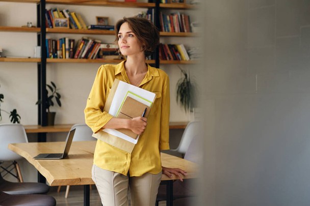 Νεαρή όμορφη γυναίκα με κίτρινο πουκάμισο που γέρνει στο γραφείο με σημειωματάριο και χαρτιά στο χέρι προσεκτικά κοιτάζοντας πέρα από το σύγχρονο γραφείο - Φωτογραφία, εικόνα