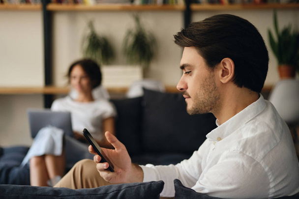 Vista lateral do jovem empresário bonito sentado no sofá com colega no fundo sonhador usando celular no espaço de co-trabalho moderno
 - Foto, Imagem