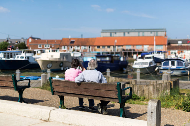 Dvojice středního věku, sedící na lavičce, obdivující řadu různých člunů kotvících v řece - Fotografie, Obrázek