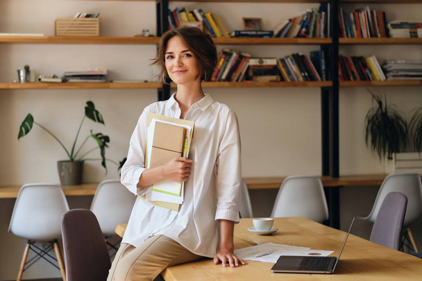Young attractive woman in white shirt happily looking in camera sitting on desk with papers and laptop in modern office - Фото, зображення