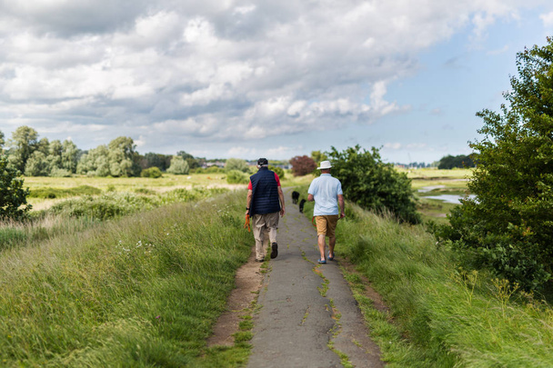 2 чоловіки середнього віку гуляють своїми собаками по популярній річковій стежці
 - Фото, зображення