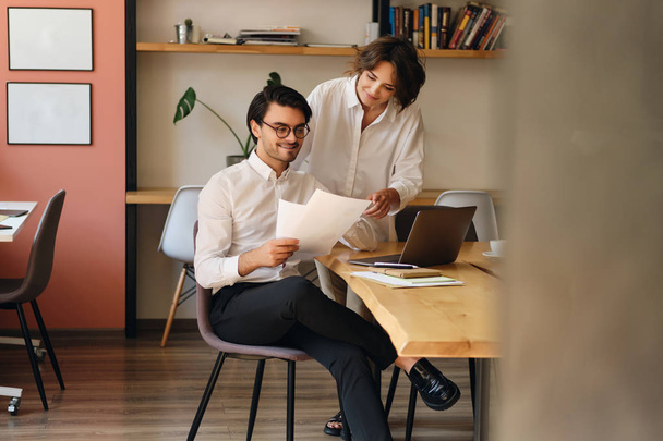 Молодые улыбающиеся деловые коллеги счастливо работают с бумагами в современном офисе
 - Фото, изображение