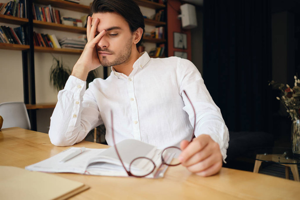 メモ帳と眼鏡をかけてテーブルに座っている若い疲れた男は、現代のオフィスで仕事で頭を疲れ果てて傾いている  - 写真・画像