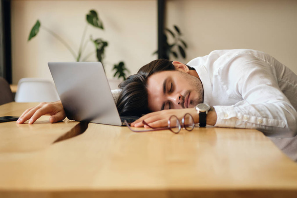 Modern ofiste iş yerinde masada uyuyan dizüstü bilgisayar ile genç yorgun adam  - Fotoğraf, Görsel
