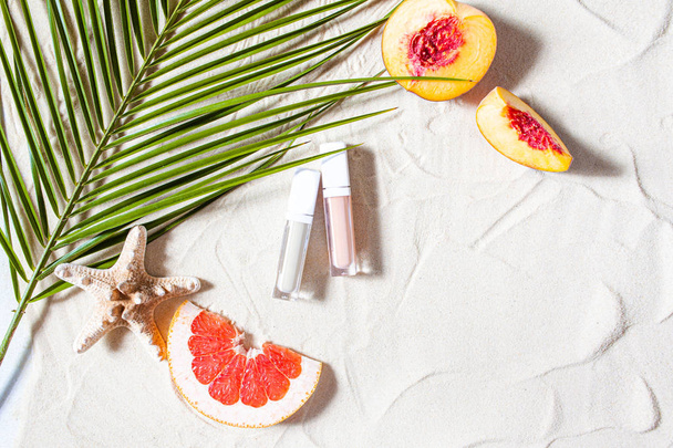 Vacation. Lip gloss in a transparent tube lies on the sand, surrounded by vibrant fruits, corals, and palm leaves. - Foto, afbeelding