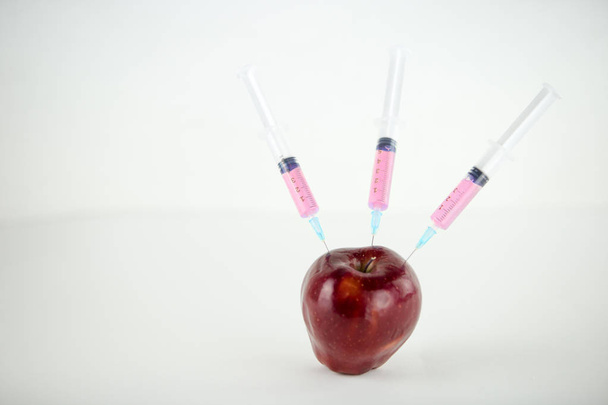 Conceito: manipulação humana de OGM da natureza e frutos envenenados relativos. Close-up de uma maçã contaminada com três seringas roscadas em fundo branco
 - Foto, Imagem