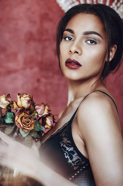 Portrait of beautiful young woman mixed race in seductive lingerie holding bouquet of dried roses. - Фото, зображення