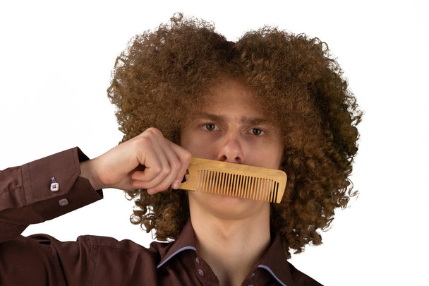 Portrait of a curly-haired young man with a wooden comb near his mouth with magnificent hair on a white isolated background. male hair care concept - Фото, зображення