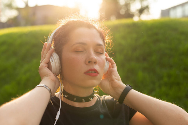 Young beautiful woman listening to relaxing music using headphones and online radio while walking through the park on a warm summer evening. Musical subscription concept. - Foto, Imagem