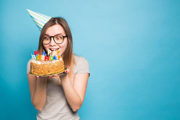 Crazy funny girl in a paper hat and glasses holding a big birthday cake on the blue background with copy space. Concept of prank and greetings. - Φωτογραφία, εικόνα