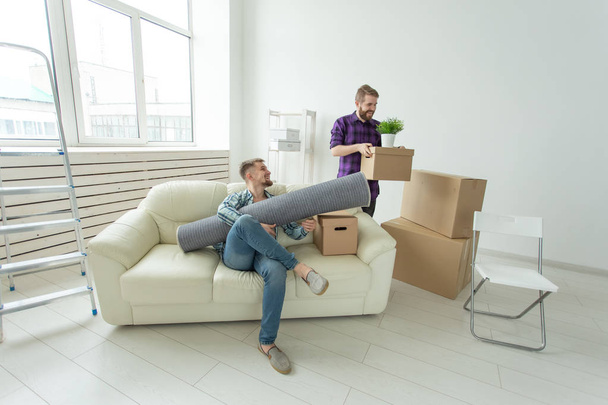 Dois estudantes amigos do sexo masculino segurando suas coisas em suas mãos sentados na sala de estar de um novo apartamento. Conceito de aquecimento doméstico
 - Foto, Imagem