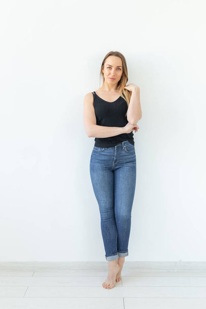Styl a lidi koncept-mladá žena v džínách, která stojí nad bílou stěnou a vypadá jako sexy - Fotografie, Obrázek