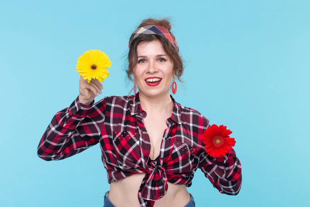 Urocza młoda kobieta uśmiechnięta trzyma dwa czerwone i pomarańczowe kwiaty w dłoniach stwarzających na niebieskim tle. Koncepcja piękna i samoopieki. - Zdjęcie, obraz