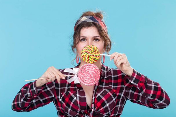 Funny beautiful young woman with bulging eyes closes her mouth with colorful lollipops posing on a blue background. Love concept and high-calorie desserts. - Foto, Bild