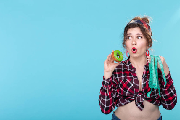 Close-up geschokt verrast jonge mooie vrouw kijken naar het meten van tape en het houden van een groene donut in handen poseren op een blauwe achtergrond copyspace. Het concept van voeding en afwijzing van schadelijke - Foto, afbeelding