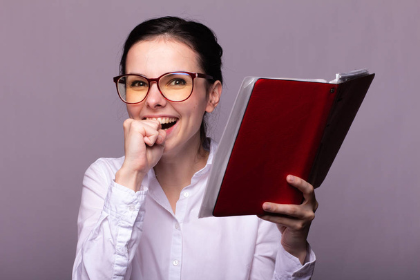 menina em uma camisa branca e óculos detém um caderno vermelho em sua mão
 - Foto, Imagem