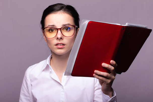 girl in a white shirt and glasses holds a red notebook in her hand - Fotoğraf, Görsel