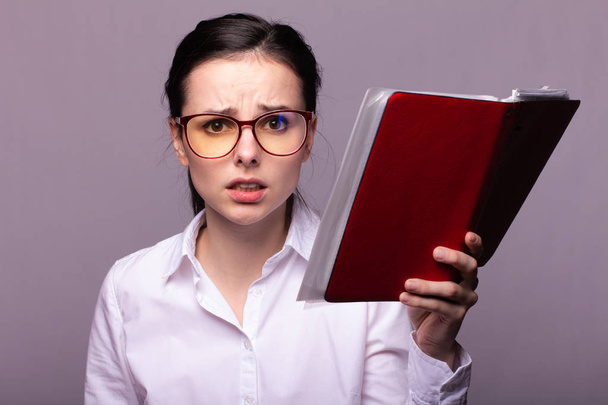 girl in a white shirt and glasses holds a red notebook in her hand - Фото, изображение