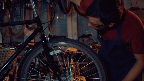 theme small business bike repair. Young brunette Caucasian man in protective glasses, gloves and fartuhe uses hand tools to repair and tuning Rim Brakes and wheel spinning bike in a garage workshop - Photo, Image