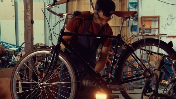 Téma kisvállalkozások kerékpár javítás. A kaukázusi barna fiatalember visel, biztonsági szemüveg, a kesztyűt és a kötényt ragaszkodik ahhoz, hogy a sebesség kapcsolók a kerékpár kormányon, a garázs, műhely - Fotó, kép