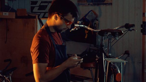 тема малый бизнес ремонт велосипедов. Юный кавказский брюнетка мужчина в защитных очках, перчатках и фартуке использует мобильные телефоны технологии, делает заметки, контрольный лист в велосипедной мастерской
 - Фото, изображение