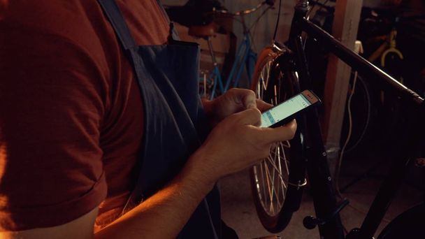 Tema küçük işletme bisiklet onarımı. Koruyucu gözlük, eldiven ve fartukhe giyen genç beyaz esmer adam cep telefonu teknolojisini kullanır, notlar, Bisiklet atölyesinde denetim listesi alır - Fotoğraf, Görsel