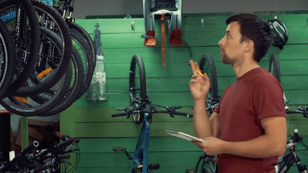 Thema Kleinunternehmen, die Fahrräder verkaufen. junge kaukasische männliche Brünette Kleinunternehmer, Filialleiter verwendet Notizblock und Stift macht Notizen, Checkliste im Fahrradgeschäft - Foto, Bild