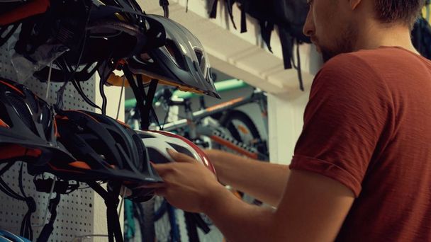 Un joven caucásico se para cerca de un puesto en una tienda de bicicletas en el departamento de protección de la cabeza. Elegir un casco de bicicleta en una pequeña tienda
 - Foto, imagen
