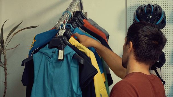 A young Caucasian man is standing near a racks of sports cycling clothes on a hanger in a bicycle store. Choosing a bicycle clothes and helmet - Photo, Image