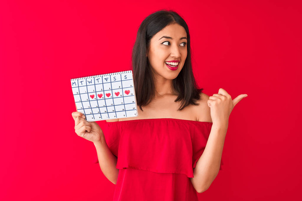 junge schöne Chinesin, die einen Kalender in der Hand hält, der über einem isolierten roten Hintergrund steht und mit dem Daumen zur Seite zeigt, mit einem glücklichen Lächeln im Gesicht - Foto, Bild