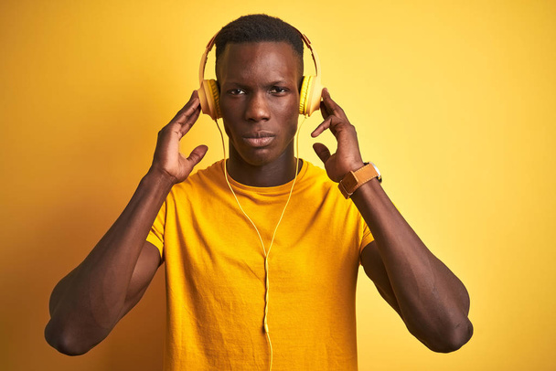 孤立した黄色の背景の上にヘッドフォンを使用して音楽を聴くアフリカ系アメリカ人男性は、真剣に考えるスマートな顔に自信を持った表情で - 写真・画像