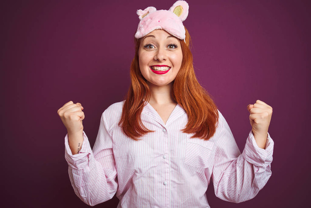 Young beautiful redhead woman wearing pajama and mask over purple isolated background celebrating surprised and amazed for success with arms raised and open eyes. Winner concept. - Photo, Image