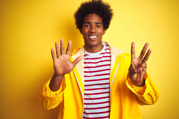 African american man with afro hair wearing rain coat standing over isolated yellow background showing and pointing up with fingers number eight while smiling confident and happy. - Фото, изображение