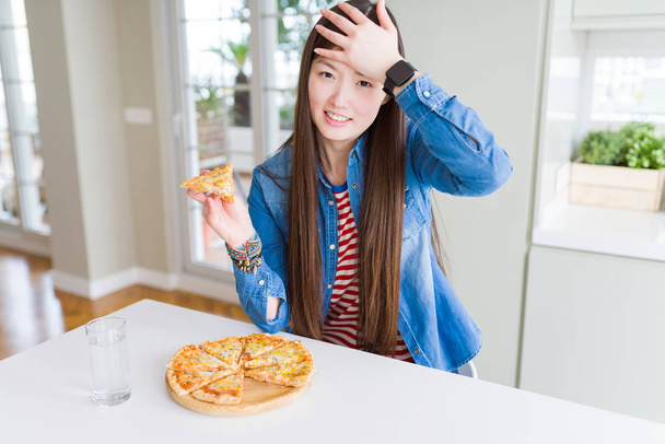 Beautiful Asian woman eating a slice of cheese pizza stressed with hand on head, shocked with shame and surprise face, angry and frustrated. Fear and upset for mistake. - Foto, Imagen