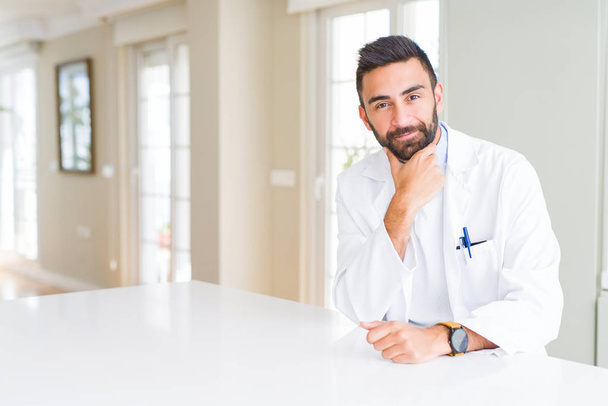 Handsome hispanic doctor or therapist man wearing medical coat at the clinic looking confident at the camera with smile with crossed arms and hand raised on chin. Thinking positive. - Photo, Image