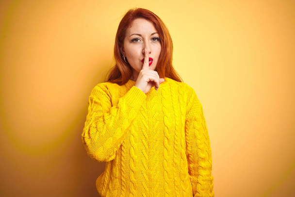 Krásná žena v zimním svetru, která stojí na izolovaném žlutém pozadí a prosí, aby byla zticha prstem na rtech. Ticho a tajný koncept. - Fotografie, Obrázek