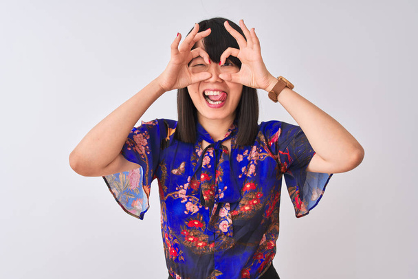 Nuori kaunis kiinalainen nainen yllään kesän kukka t-paita yli eristetty valkoinen tausta tekee ok ele kuin kiikarit kiinni kielen ulos, silmät katsellen sormien läpi. Hullu ilme
. - Valokuva, kuva