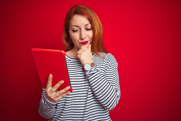 Young beautiful redhead woman using tablet over red isolated background serious face thinking about question, very confused idea - Photo, Image