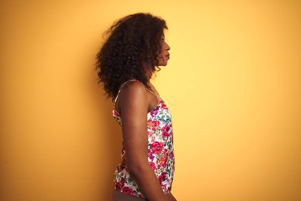 Afro-amerikai nő visel virágos nyári póló alatt elszigetelt sárga háttér látszó oldalra, Relax profil pózol természetes arcát magabiztos mosoly. - Fotó, kép