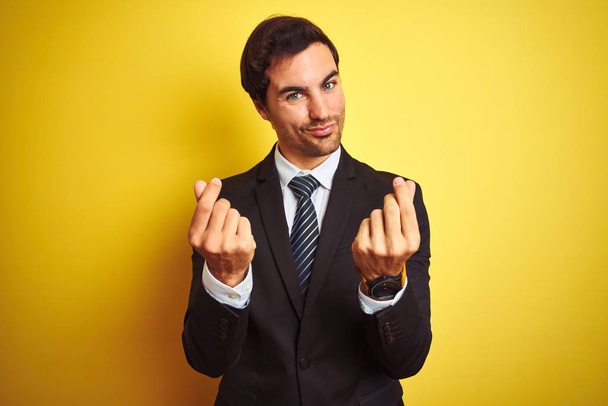 Young handsome businessman wearing suit and tie standing over isolated yellow background doing money gesture with hands, asking for salary payment, millionaire business - Photo, Image