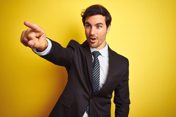 Young handsome businessman wearing suit and tie standing over isolated yellow background Pointing with finger surprised ahead, open mouth amazed expression, something on the front - Foto, Imagem