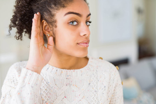 Krásná mladá africká americká žena s afro vlasy nosí ležérní svetr s úsměvem s rukou přes ucho naslouchá slyšení zvěsti nebo drby. Hluchota koncept. - Fotografie, Obrázek