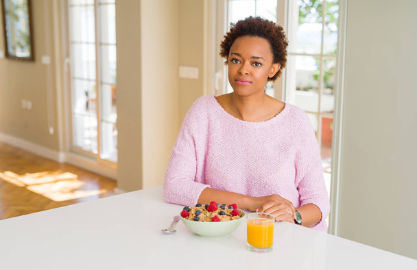 Mujer afroamericana joven desayunando saludable por la mañana en casa Relajado con expresión seria en la cara. Simple y natural mirando a la cámara
. - Foto, imagen