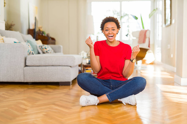 Młoda piękna African American kobieta siedzi na podłodze w domu obchodzi zaskoczony i zdumiony sukcesem z ramionami podniesione i otwarte oczy. Koncepcja zwycięzcy. - Zdjęcie, obraz