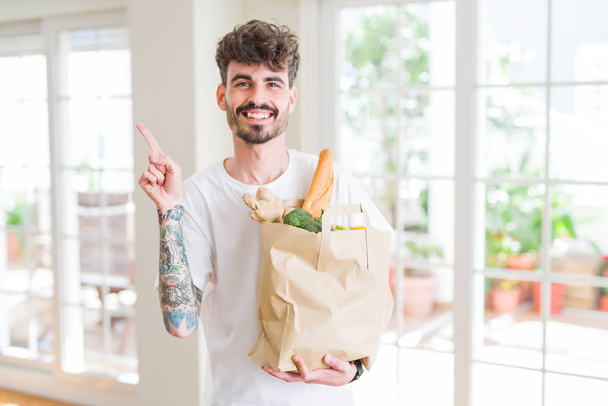 Nuori mies pitäen paperipussi tuoreita elintarvikkeita supermarketista erittäin onnellinen osoittaa käsin ja sormella sivulle
 - Valokuva, kuva